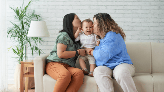 lesbian couple sitting on couch with smiling baby boy standing between them kissing either side of his face