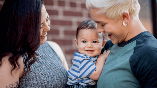lesbian moms holding infant son smiling against brick wall background