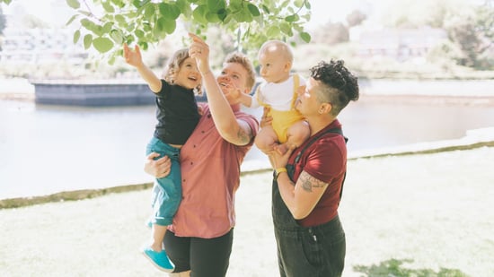 lesbian moms holding two young children pointing up at a tree outside
