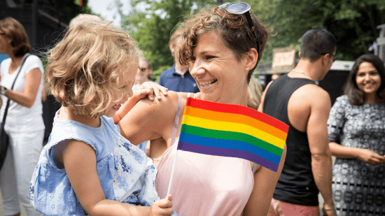 queer mother holding young daughter waving pride flag at parade